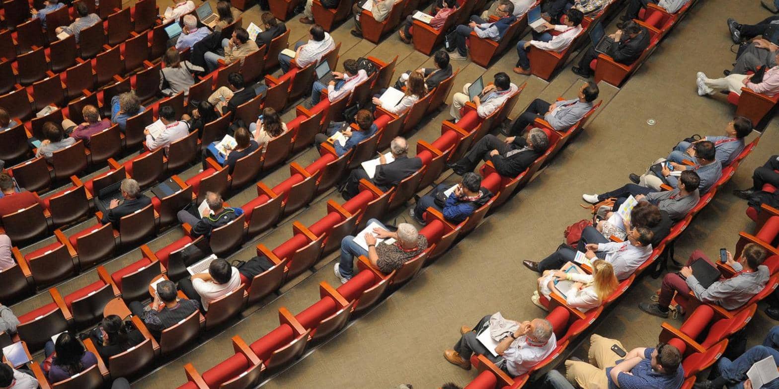 Overhead view of a conference hall at COP26 with attendees seated in red chairs, some taking notes while listening to a presentation on climate change.