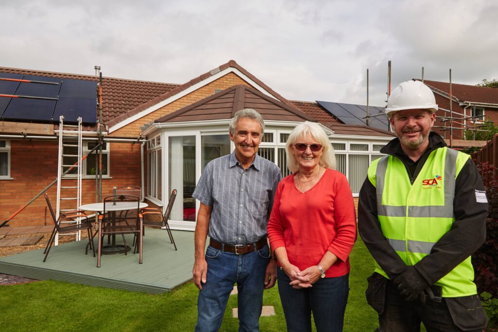 An elderly couple standing with a domestic solar installer outside their home, which has solar panels on the roof installed by scasolar team.