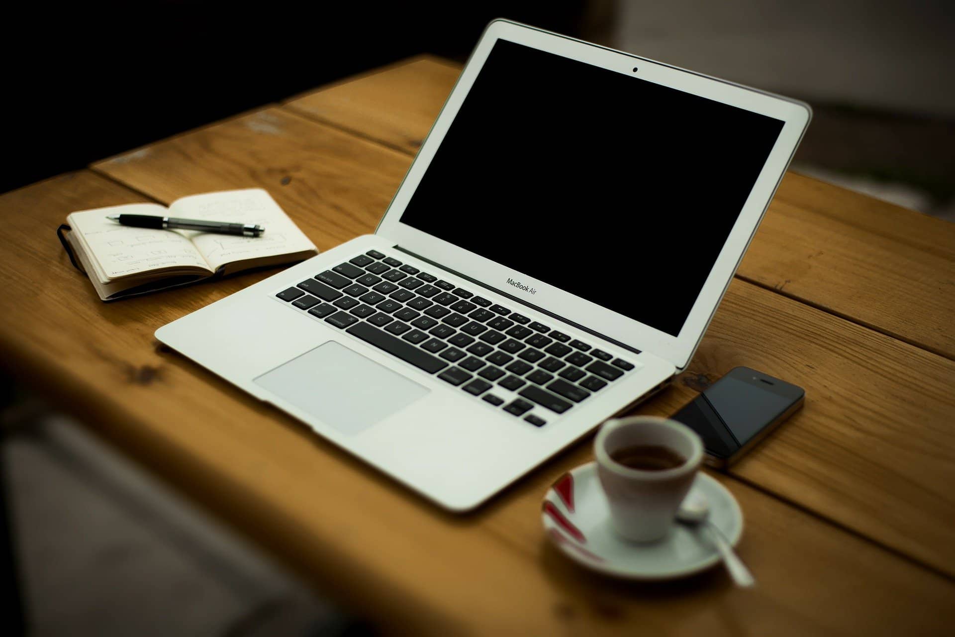 A laptop with a black screen, a smartphone, a blog template notebook with a pen, and a cup of coffee on a wooden table.