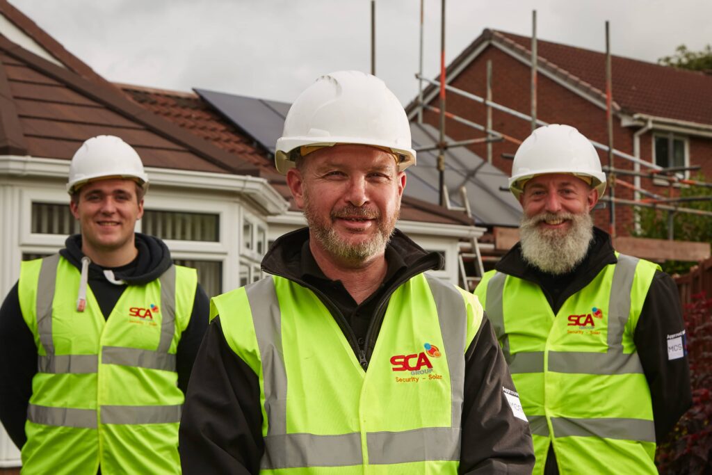 Three SCA Solar installers from a solar panel installation company in Cheshire, in safety gear, including hard hats and reflective vests, smiling at the camera with a residential area in the background.