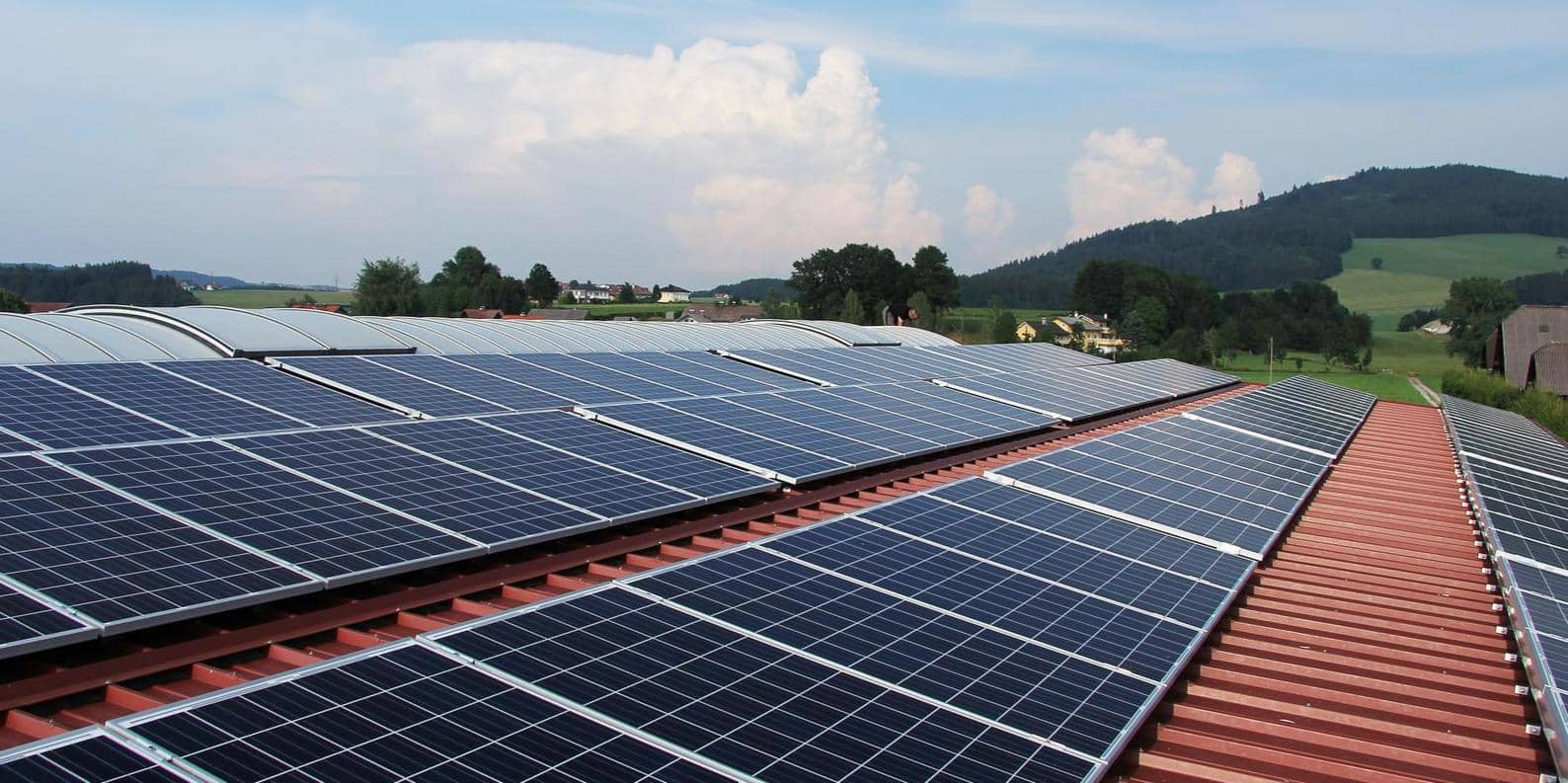 Solar panels installed on the roof of a commercial building in a rural setting with green hills in the background and a clear sky.