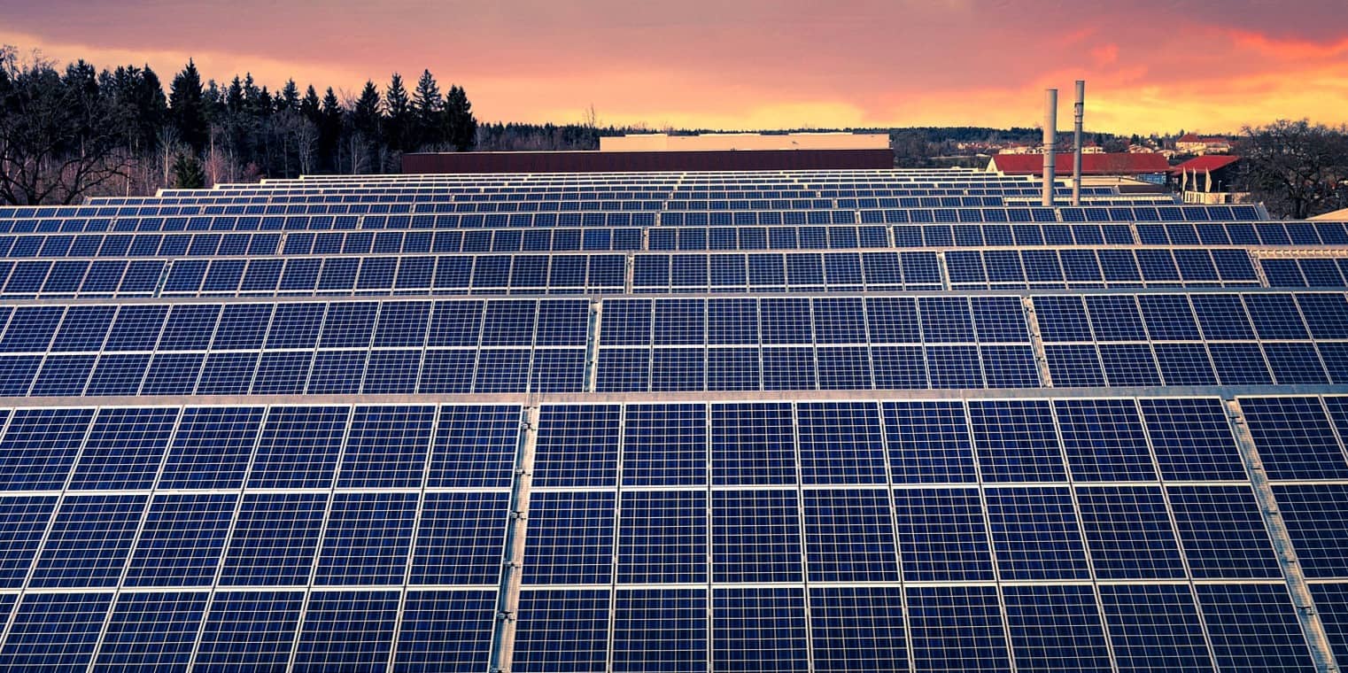 Solar panels make electricity on a roof against a sunset sky with trees and distant buildings in the background.