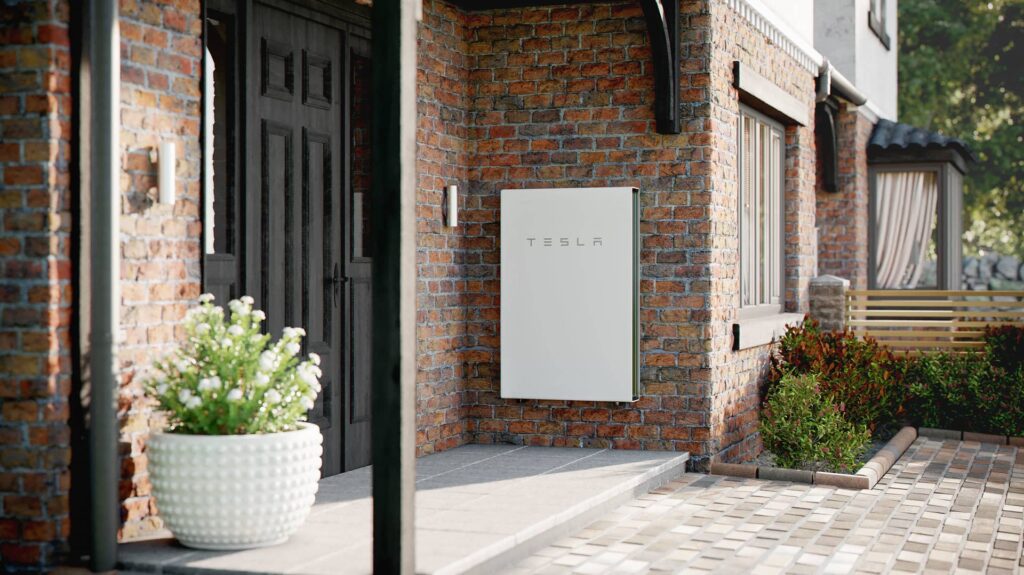 A Tesla Powerwall solar battery storage mounted on the brick exterior wall of a residential home near the entrance.