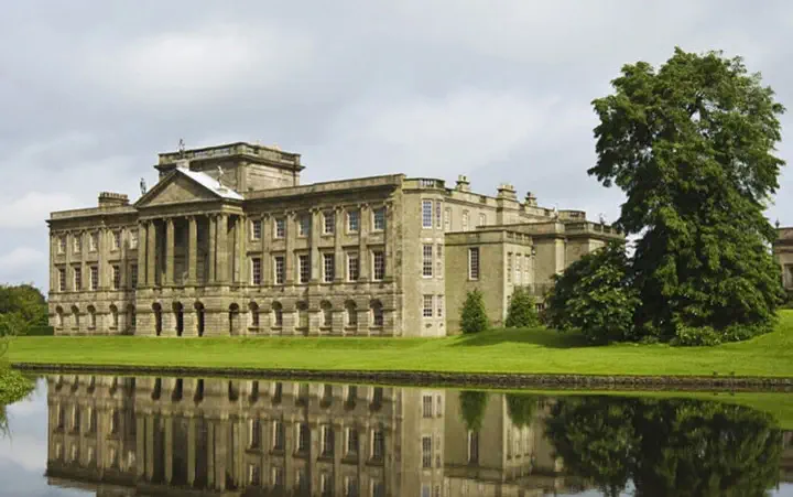 A large classical mansion with extensive columns and sculptures, reflected in a still pond, surrounded by lush green trees.