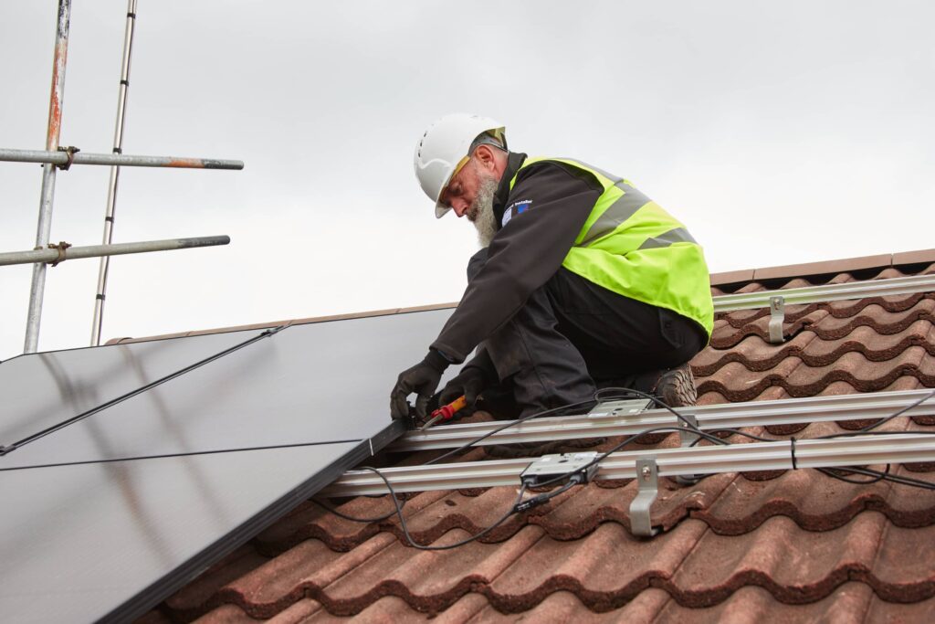 A worker in a helmet and reflective vest installs solar panels with micro inverters on a tiled roof.