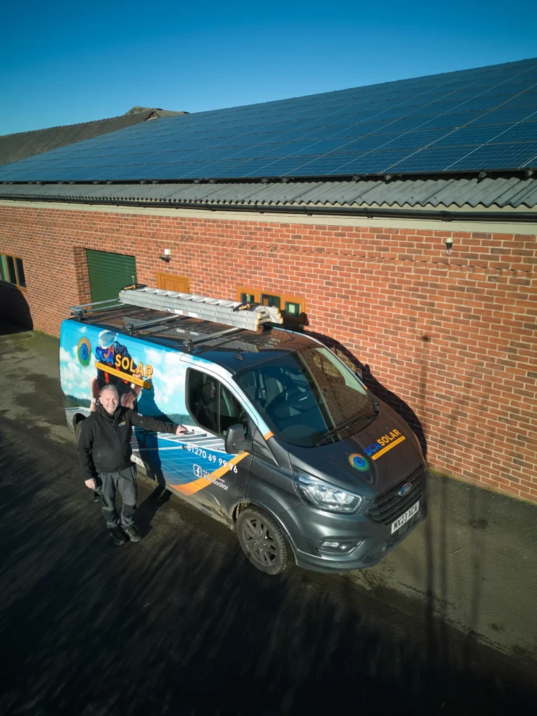 A scasolar installer stands beside a decorated scasolar company van with solar panels on the building roof in the background under a clear sky.