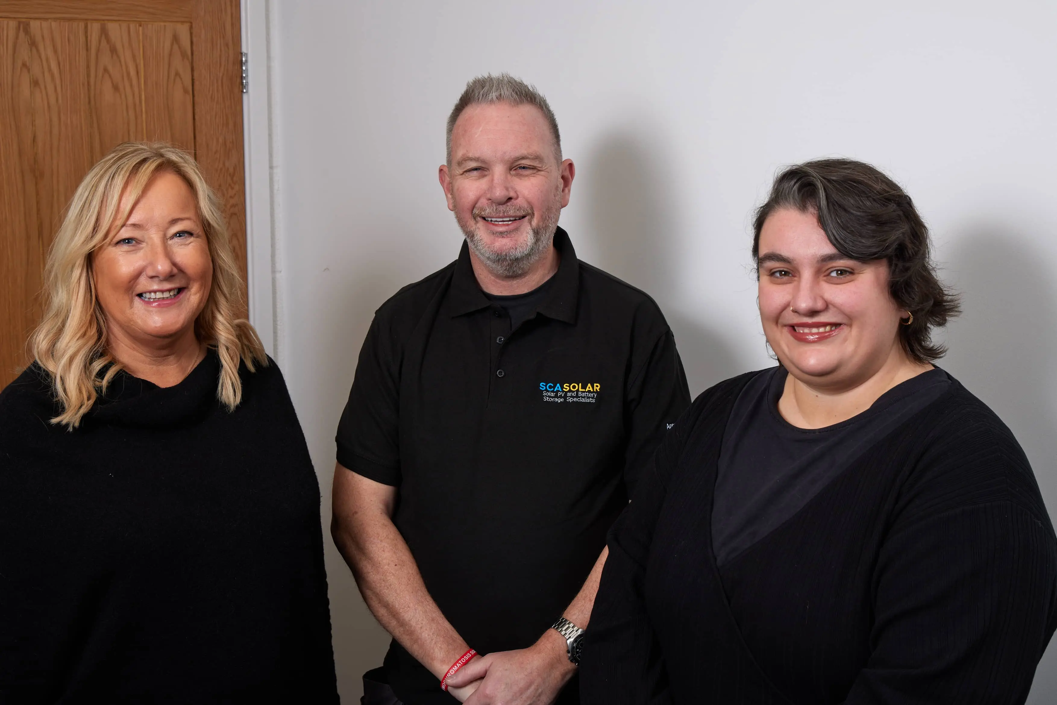 Three Professional Solar Panel Installers based in Cheshire standing and smiling indoors, two women and one man in the middle wearing a black shirt with "SCASOLAR" logo.