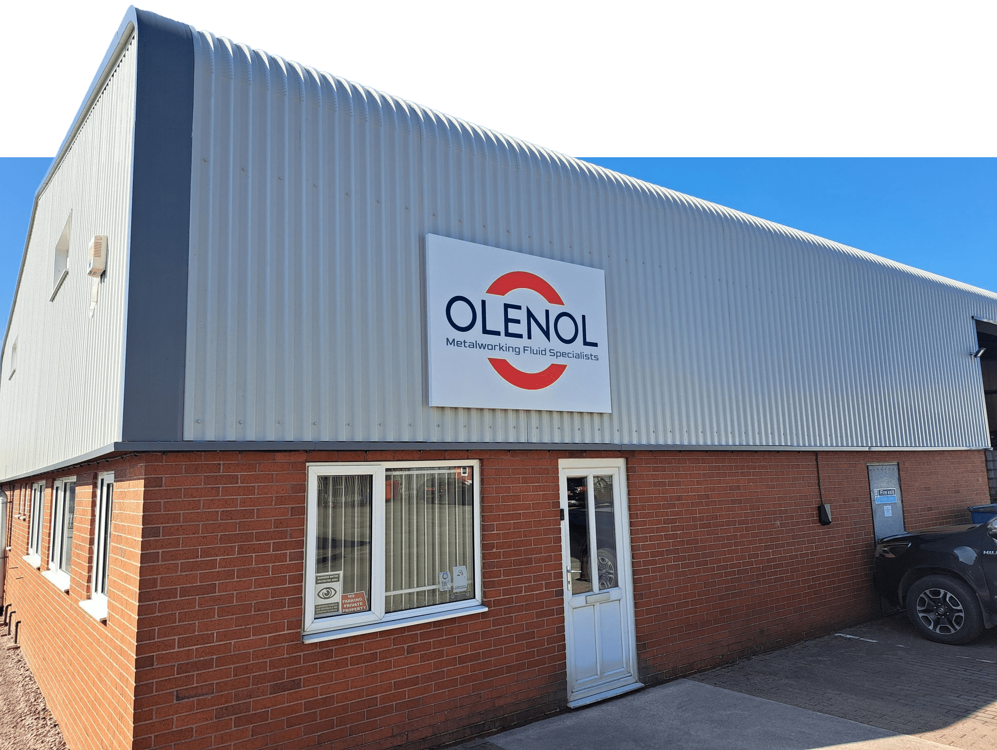 A modern industrial building with a brick lower half and metal siding, displaying a sign that reads "OLENOL Metalworking Fluid Specialists" above the entrance—designed to future-proof operations and reduce carbon footprint.
